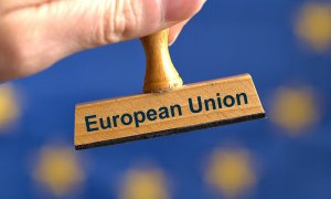 Un sello de madera simbólico con la inscripción "Unión Europea", sostenido por una mano frente a una bandera borrosa de la UE en el fondo.