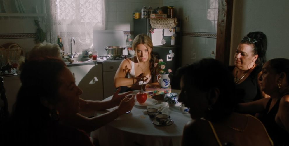 Lola Índigo sentada en una mesa de cocina rodeada de mujeres. La imagen es un fotograma de su videoclip 'Mala Suerte'.